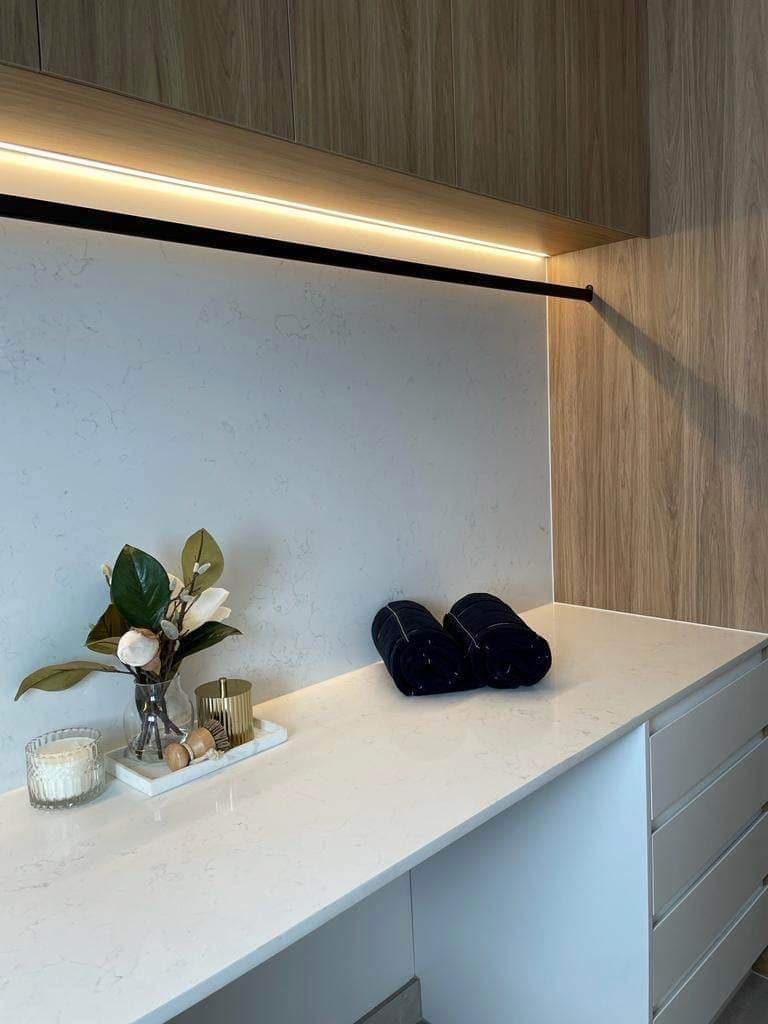 Strip lighting beneath timber cabinets above a white bench and tiled splashback